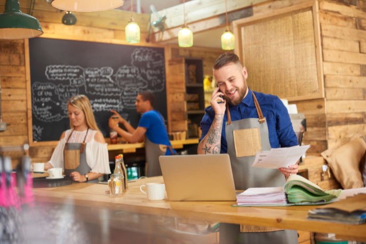 A small business owner wearing an apron smiles while talking on the phone and reviewing documents at a rustic cafe counter