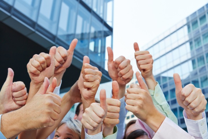 A group of people giving a collective thumbs up gesture in front of a modern office building