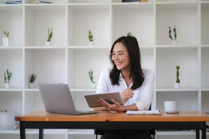 A businesswoman wearing glasses works happily on her laptop and tablet